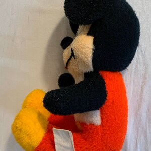Puede incluir: Un juguete de peluche vintage de Mickey Mouse con pelaje negro, pantalones rojos y amarillos y una etiqueta blanca en el lateral.
