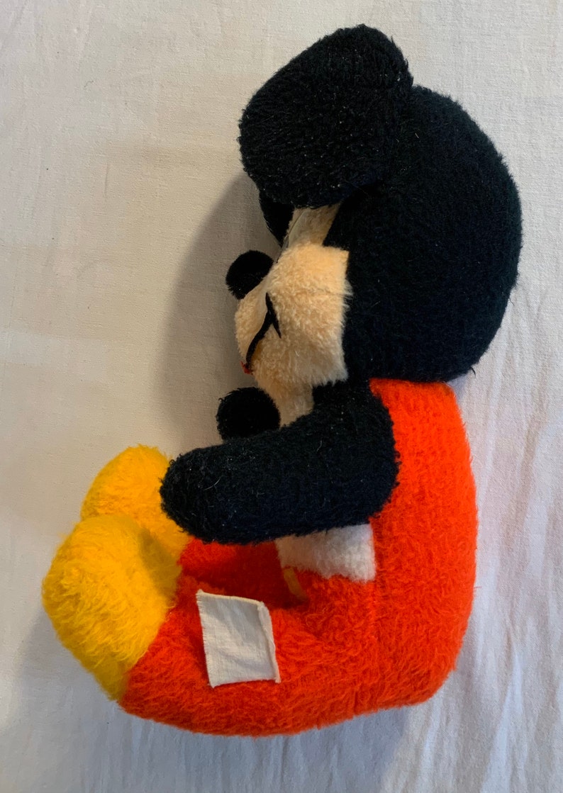 Puede incluir: Un juguete de peluche vintage de Mickey Mouse con pelaje negro, pantalones rojos y amarillos y una etiqueta blanca en el lateral.