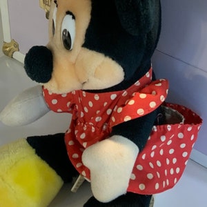 May include: A plush toy of a cartoon mouse wearing a red dress with white polka dots and a red bow. The mouse has black ears, a black nose, and yellow shoes.
