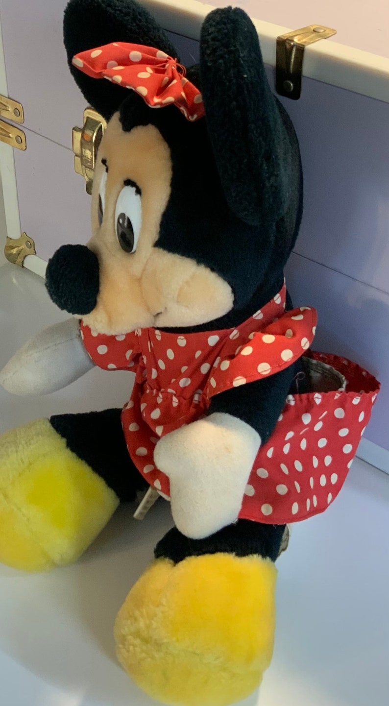 May include: A plush toy of a cartoon mouse wearing a red dress with white polka dots and a red bow. The mouse has black ears, a black nose, and yellow shoes.