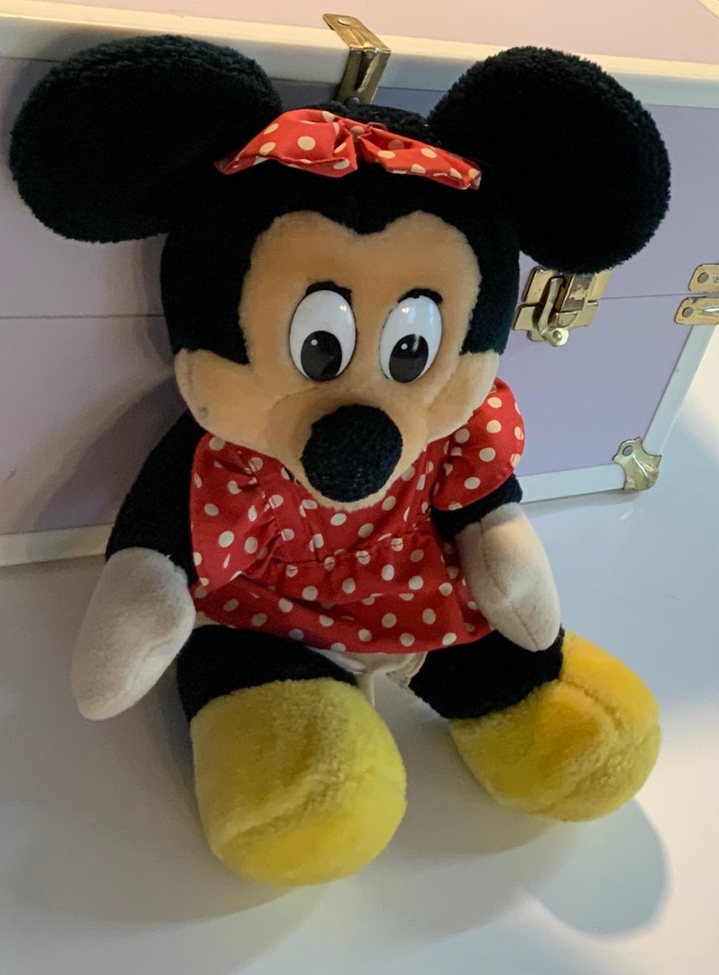 May include: A plush toy of Mickey Mouse's girlfriend Minnie Mouse. She is wearing a red dress with white polka dots and a red bow with white polka dots. She has large black eyes and a black nose.