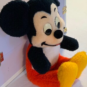 Puede incluir: Un juguete de peluche de Mickey Mouse con orejas negras, un cuerpo negro, pantalones rojos y zapatos amarillos. El juguete est&aacute; sentado sobre una superficie blanca.