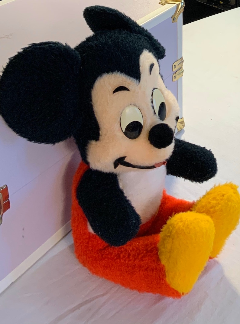 Puede incluir: Un juguete de peluche de Mickey Mouse con orejas negras, un cuerpo negro, pantalones rojos y zapatos amarillos. El juguete est&aacute; sentado sobre una superficie blanca.