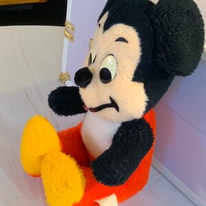 Puede incluir: Un juguete de peluche de Mickey Mouse sentado con las piernas cruzadas. El juguete lleva pantalones rojos con zapatos amarillos y tiene orejas negras y una nariz negra.