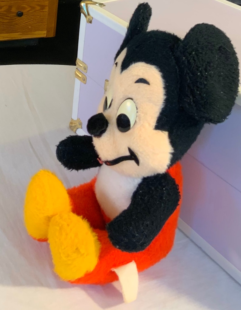 Puede incluir: Un juguete de peluche de Mickey Mouse sentado con las piernas cruzadas. El juguete lleva pantalones rojos con zapatos amarillos y tiene orejas negras y una nariz negra.