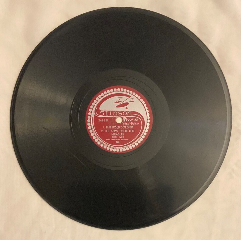 May include: A black vinyl record with a red label. The label has a white border and the text "Stinson Records Vocal-Guitar 345-1-B 1. THE BOLD SOLDIER 2. THE SOW TOOK THE MEASLES BURL IVES The Singing Cowboy"