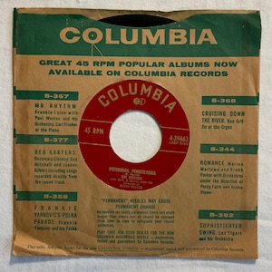 May include: A vintage Columbia Records advertisement featuring a green and white label with a red center. The label has the Columbia Records logo and the text "Great 45 RPM Popular Albums Now Available on Columbia Records". The label also features a list of popular albums with their corresponding record numbers and titles. The text "Play safe. Ask your dealer for the new Columbia Needle - engineered, tested and guaranteed by Columbia Records." is at the bottom of the label.