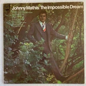 May include: A vintage vinyl record album cover featuring a man in a suit standing in a woodland setting. The album cover is titled "Johnny Mathis/The Impossible Dream" and lists the songs "Strangers in the Night", "On a Clear Day You Can See Forever", "Somewhere, My Love", "I Will Wait for You", "So Nice", "Go Away Little Girl", "Eleanor Rigby", "The Very Thought of You", "Moment to Moment", and "The Impossible Dream".