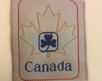 May include: A white fabric label with a red stitched border. Inside the border is a gold outline of a maple leaf with a blue shamrock in the centre. The word "Canada" is written in blue below the leaf.