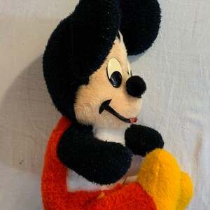 Puede incluir: Un juguete de peluche de Mickey Mouse sentado con las piernas cruzadas. El juguete lleva pantalones rojos con detalles amarillos y tiene pelo negro con la cara de color canela.