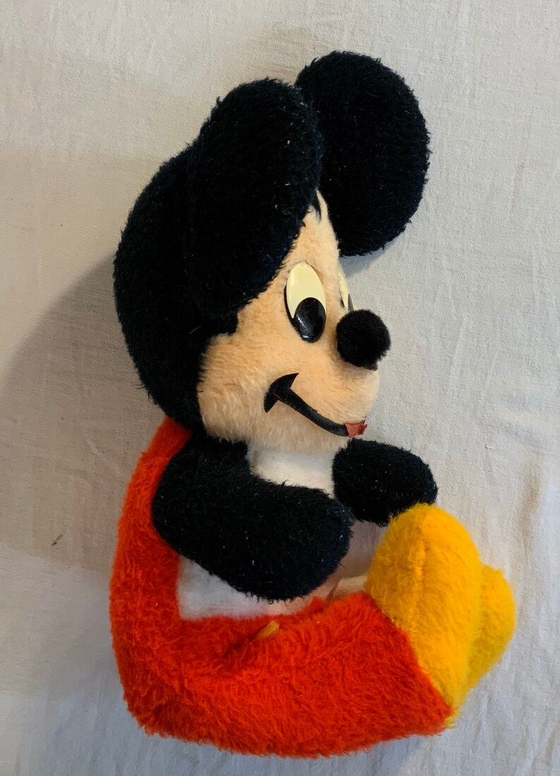 Puede incluir: Un juguete de peluche de Mickey Mouse sentado con las piernas cruzadas. El juguete lleva pantalones rojos con detalles amarillos y tiene pelo negro con la cara de color canela.
