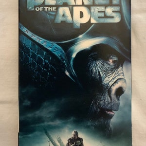 Pode incluir: Uma capa de VHS para o filme "O Planeta dos Macacos" com Mark Wahlberg. A capa apresenta um close-up de um macaco de pele escura com uma expressão séria. O título do filme está em letras brancas grandes sobre um fundo azul.