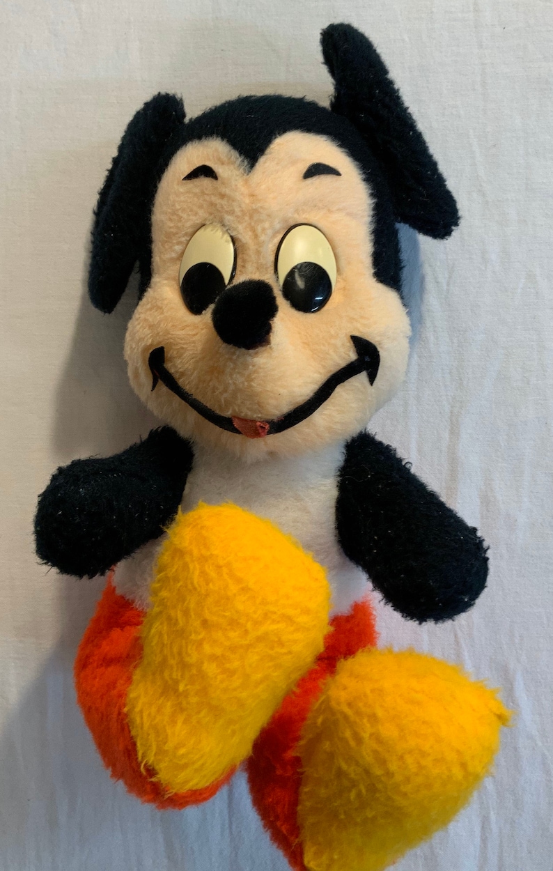 Puede incluir: Un juguete de peluche vintage de Mickey Mouse con orejas negras, brazos negros y zapatos amarillos y naranjas. El juguete tiene una cara blanca con ojos negros y una lengua roja que sobresale.