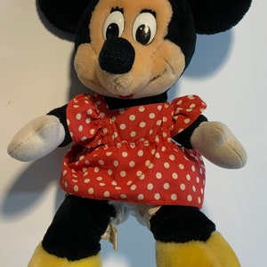 May include: A plush Minnie Mouse toy wearing a red dress with white polka dots and a red bow. The toy has black ears, a black nose, and yellow shoes.
