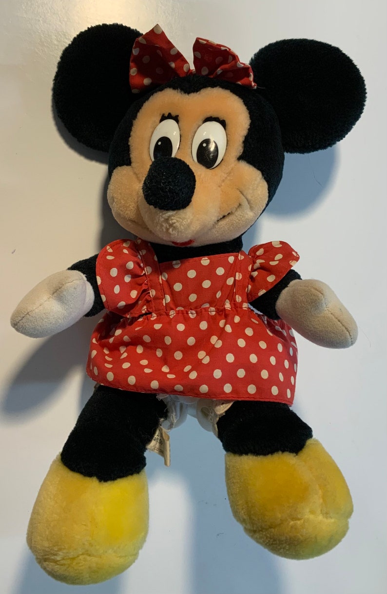 May include: A plush Minnie Mouse toy wearing a red dress with white polka dots and a red bow. The toy has black ears, a black nose, and yellow shoes.