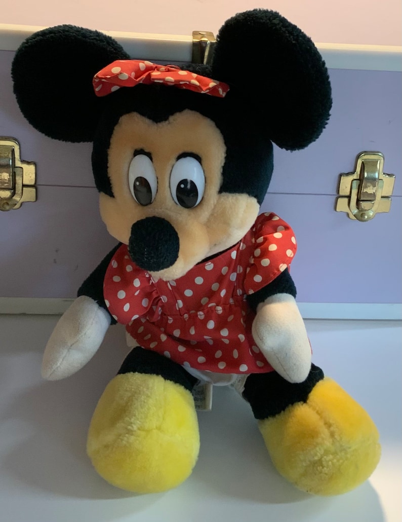 May include: A plush toy of Minnie Mouse wearing a red dress with white polka dots and a red bow. She has black ears, a black nose, and yellow shoes.