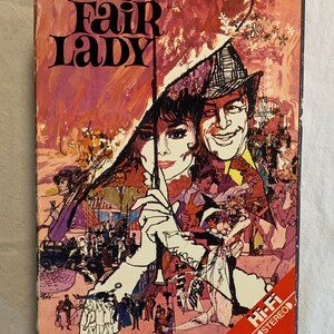 May include: A VHS tape cover for the movie "My Fair Lady" featuring Audrey Hepburn and Rex Harrison. The cover is a vibrant mix of red, orange, yellow, and pink with a stylized illustration of the actors.