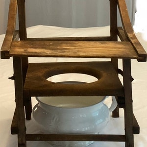 Może przedstawiać: Antyczne drewniane krzesło komodowe z białym porcelanowym nocnikiem. Krzesło ma drewniane siedzisko z otworem w środku i oparcie. Krzesło jest wykonane z ciemnobrązowego drewna i ma rustykalny wygląd.