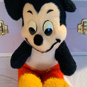 Puede incluir: Un juguete de peluche vintage de Mickey Mouse con pelaje negro, orejas grandes y una cara blanca. Lleva pantalones cortos rojos con zapatos amarillos y tiene una gran sonrisa.