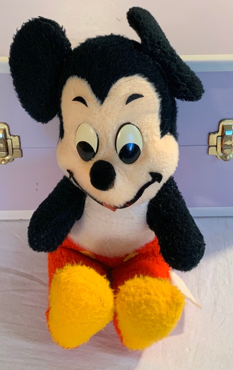 Puede incluir: Un juguete de peluche vintage de Mickey Mouse con pelaje negro, orejas grandes y una cara blanca. Lleva pantalones cortos rojos con zapatos amarillos y tiene una gran sonrisa.