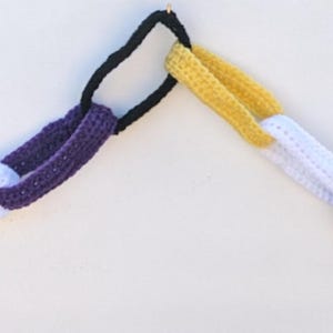 May include: A purple, white and yellow crocheted chain garland. The chain is made of individual loops of yarn, each with a different colour.