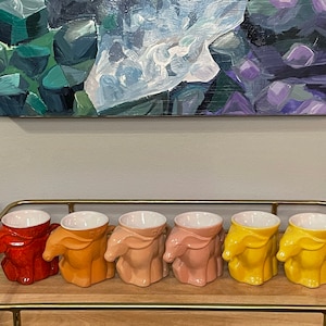 May include: Six ceramic mugs shaped like horses in a gradient of colors from orange to yellow. The mugs are arranged in a row on a wooden surface.