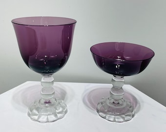 May include: Two elegant, stemmed glasses. The bowls are a deep purple, transitioning to a lighter shade at the rim. The stems are clear, with a decorative, rounded design. The bases are clear, with a scalloped edge. Perfect for serving beverages.