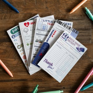 May include: A collection of stationery items, including pens and printed papers, arranged on a wooden surface. The papers feature text such as "Baby", "Pet", "Little Travelers", "Boarding Pass", and "Rock Shop Receipt". Pens are in various colors.