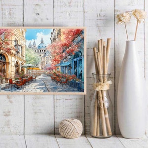May include: A watercolor painting of a Parisian street scene with a cafe and tables and chairs. The buildings are painted in a light yellow color with red tile roofs. The street is lined with trees with pink and red flowers. The sky is blue with white clouds.