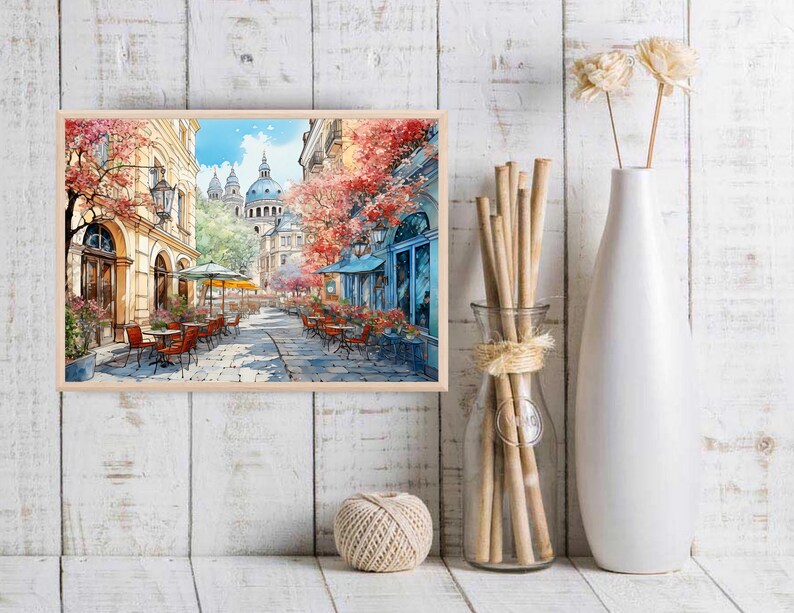 May include: A watercolor painting of a Parisian street scene with a cafe and tables and chairs. The buildings are painted in a light yellow color with red tile roofs. The street is lined with trees with pink and red flowers. The sky is blue with white clouds.