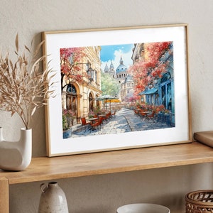 May include: A framed watercolor painting of a European street scene with a cafe and buildings. The cafe has tables and chairs set up outside, and there are trees and flowers in bloom. The buildings are painted in a light yellow color, and the sky is a bright blue.