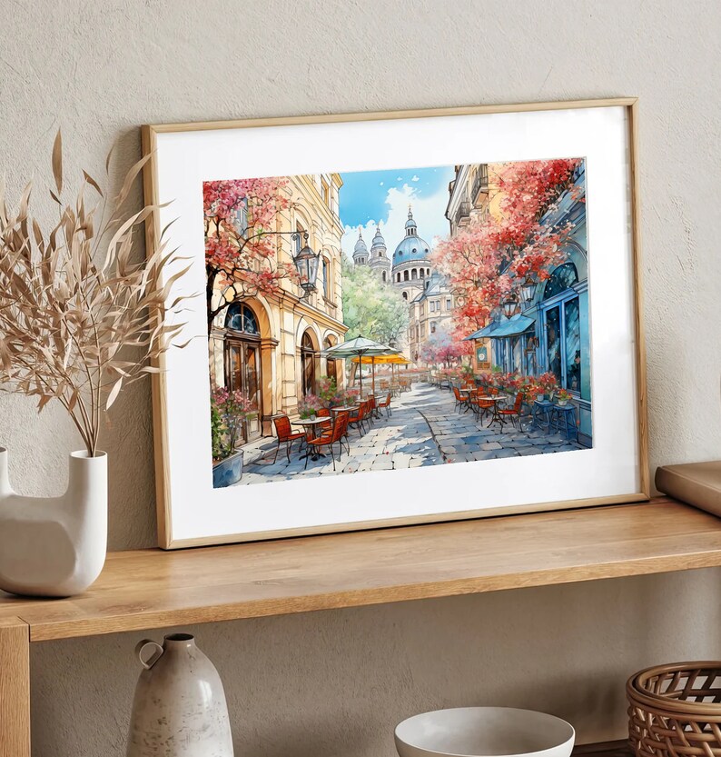 May include: A framed watercolor painting of a European street scene with a cafe and buildings. The cafe has tables and chairs set up outside, and there are trees and flowers in bloom. The buildings are painted in a light yellow color, and the sky is a bright blue.