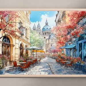 May include: A watercolor painting of a European street scene with a cafe on a cobblestone street. The cafe has tables and chairs with red umbrellas. There are buildings in the background with a large dome structure. The buildings are painted in a light yellow color with pink and red flowers on the trees.
