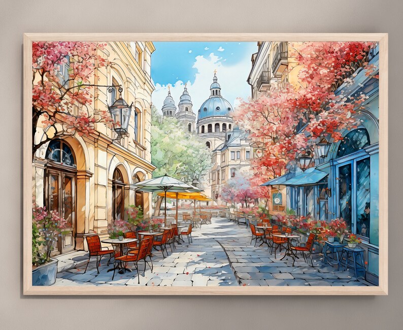May include: A watercolor painting of a European street scene with a cafe on a cobblestone street. The cafe has tables and chairs with red umbrellas. There are buildings in the background with a large dome structure. The buildings are painted in a light yellow color with pink and red flowers on the trees.