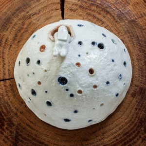 Könnte beinhalten: Weiße Keramikskulptur eines Mondes mit einer kleinen weißen Figur auf der Oberseite. Der Mond ist mit kleinen blauen und braunen Punkten bedeckt.