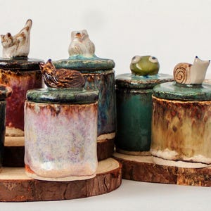 May include: A collection of seven cylindrical ceramic jars with lids, each topped with a unique sculpted figure including an octopus, cat, owl, fox, frog, snail, and mushroom. The jars feature varied glazed finishes in blue, green, red, and tan.