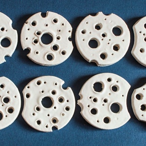 May include: Eight white ceramic coasters with a circular design featuring various sized holes. The coasters have a slightly irregular shape and a textured surface.
