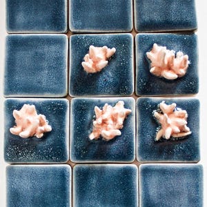May include: A grid of twelve square ceramic tiles. Nine tiles are a textured, deep blue color. Three tiles feature a raised, coral-like design in a light peach color. The tiles are arranged in a 3x4 pattern.