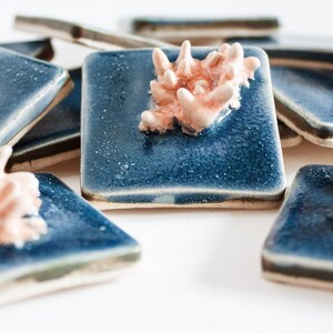 May include: A collection of square, ceramic coasters with a glossy, deep blue glaze. Each coaster features a raised, coral-like design in a soft, peachy-pink hue. The coasters are arranged on a white surface.