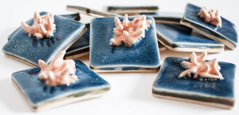May include: A collection of square, ceramic coasters with a glossy, deep blue glaze. Each coaster features a raised, coral-like design in a soft, peachy-pink hue. The coasters are arranged on a white surface.