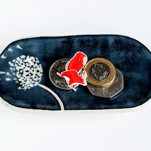 Op de afbeelding: Een donkerblauwe keramische sieradenschaal met een wit paardenbloemontwerp en een witte rand. De schaal bevat verschillende munten en een rood decoratief element. De schaal is ovaal van vorm en ligt op een wit oppervlak.