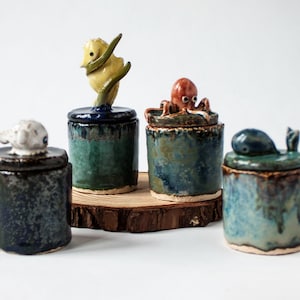 Puede incluir: Cuatro tarros de cerámica con tapas, cada uno con una figura de animal única. Los tarros tienen un acabado texturizado y vidriado en tonos de azul, verde y marrón. Uno tiene un pájaro, otro un pulpo, una ballena y una oveja.