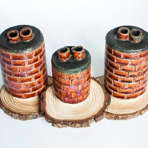 Puede incluir: Tres recipientes de cerámica con forma de chimenea con un patrón de ladrillos en tonos marrones y rojos. Cada chimenea tiene una parte superior verde oscuro con dos aberturas circulares marrones. Se exhiben sobre rodajas de madera.