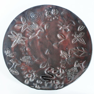 May include: A round, dark brown ceramic plate with a raised design of various nature elements, including birds, leaves, flowers, a snake, and a dragonfly. The plate has a rustic, earthy feel.