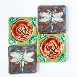 May include: Four ceramic tiles with a green background and brown and white dragonfly designs. Two tiles feature a brown circle with a stylized design inside. Two tiles feature a dragonfly with white wings and a green body.