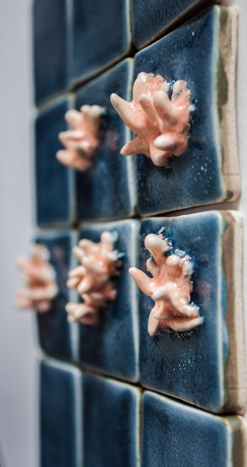 May include: Close-up of a wall installation featuring square, navy blue ceramic tiles. Each tile has a coral-like, peach-colored ceramic sculpture affixed to its surface, creating a textured, oceanic aesthetic.