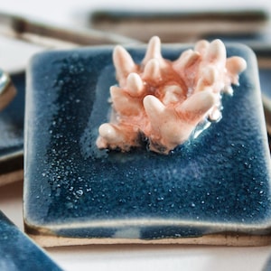 Puede incluir: Colección de baldosas de cerámica cuadradas con un esmalte azul oscuro brillante. Cada baldosa presenta una escultura en relieve con forma de coral en tonos melocotón y crema. Disposición en poca profundidad de campo.