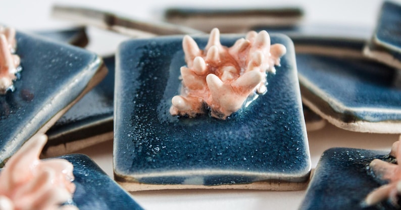 May include: A collection of square ceramic tiles with a glossy, deep blue glaze. Each tile features a raised, coral-like sculpture in shades of peach and cream. The tiles are arranged in a shallow depth of field.