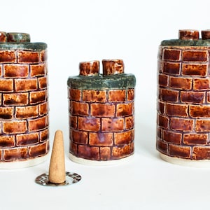 May include: Three ceramic chimney-shaped incense holders with a brick pattern in shades of brown and a green top. A cone-shaped incense stick sits on a silver disc in front of the holders. The holders have a white base.