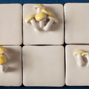 May include: Six white ceramic tiles with yellow mushroom designs. Each tile features a different arrangement of mushrooms.